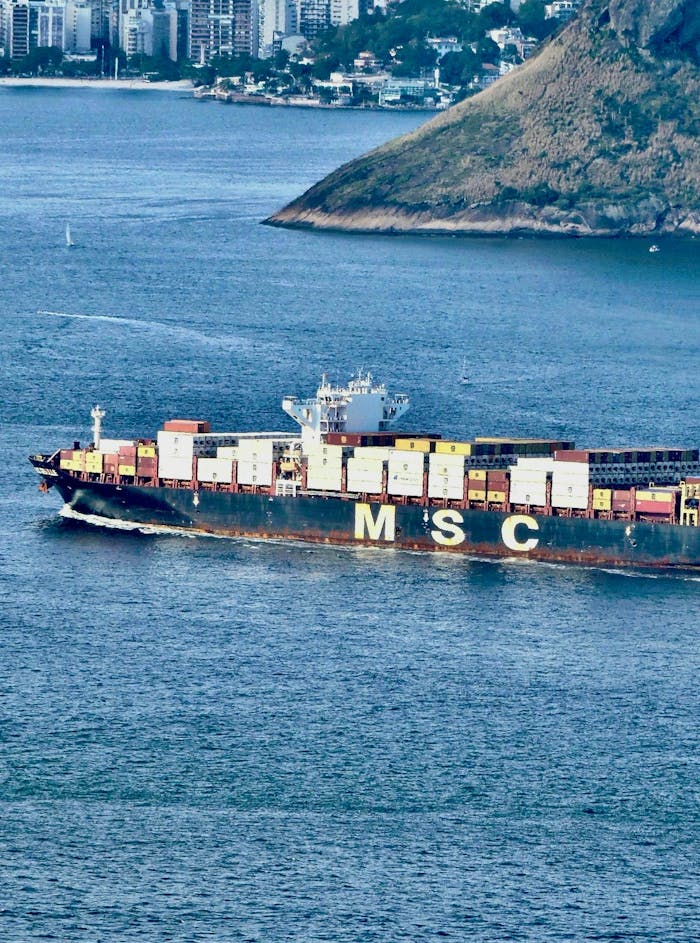 Aerial view of a massive cargo ship sailing near a coastal city with a hillside in the background.