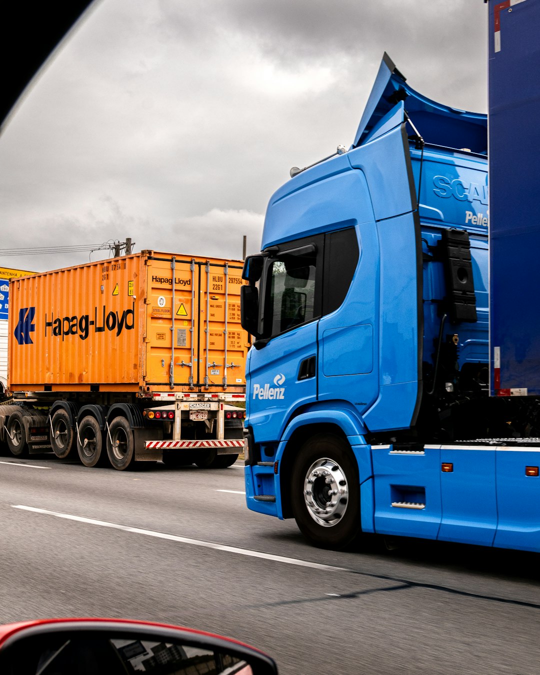 A blue Pellenz truck is driving on the highway next to an orange Hapag-Lloyd shipping container.