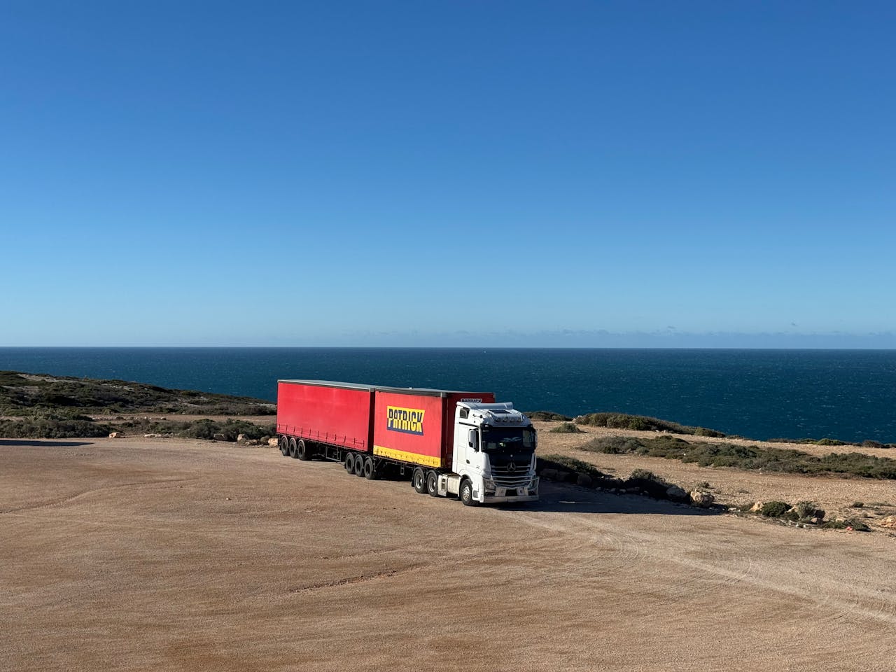 A white truck with a red trailer travels on a coastal road next to the ocean.